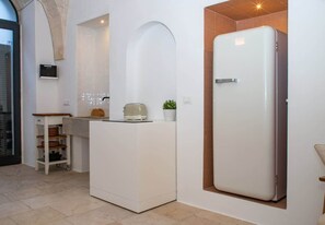 A compact kitchen with a built-in refrigerator, a small countertop, and a wooden shelf unit. - Summer - The Fab Stay (Ostuni)