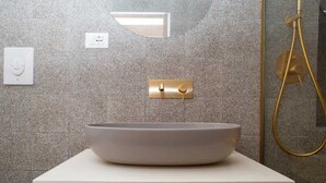 A bathroom with a large, round, white sink on a white countertop, a gold showerhead, and a wall-mounted light fixture. - Spring - The Fab Stay (Ostuni)