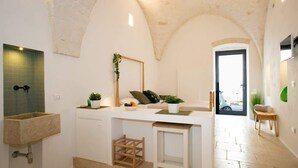 A modern kitchen with a stone sink, a wooden stool, and a view of the street through an open door. - Spring - The Fab Stay (Ostuni)