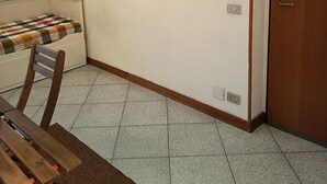 Una stanza con un letto, una sedia di legno, un tavolino, un quadro appeso e una porta.