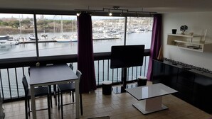 Ein modernes Zimmer mit großem Fenster mit Blick auf eine Marina. Es beinhaltet einen Esstisch, Stühle, einen Fernseher und eine Sitzecke.
