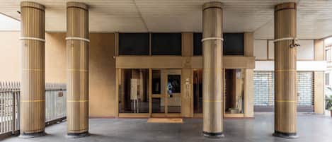 Una entrada de edificio con zona cubierta, columnas grandes y suelo texturizado.