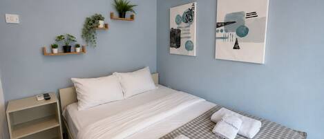A hotel room with a bed, a bedside table, a shelf with plants, and artwork on the wall.