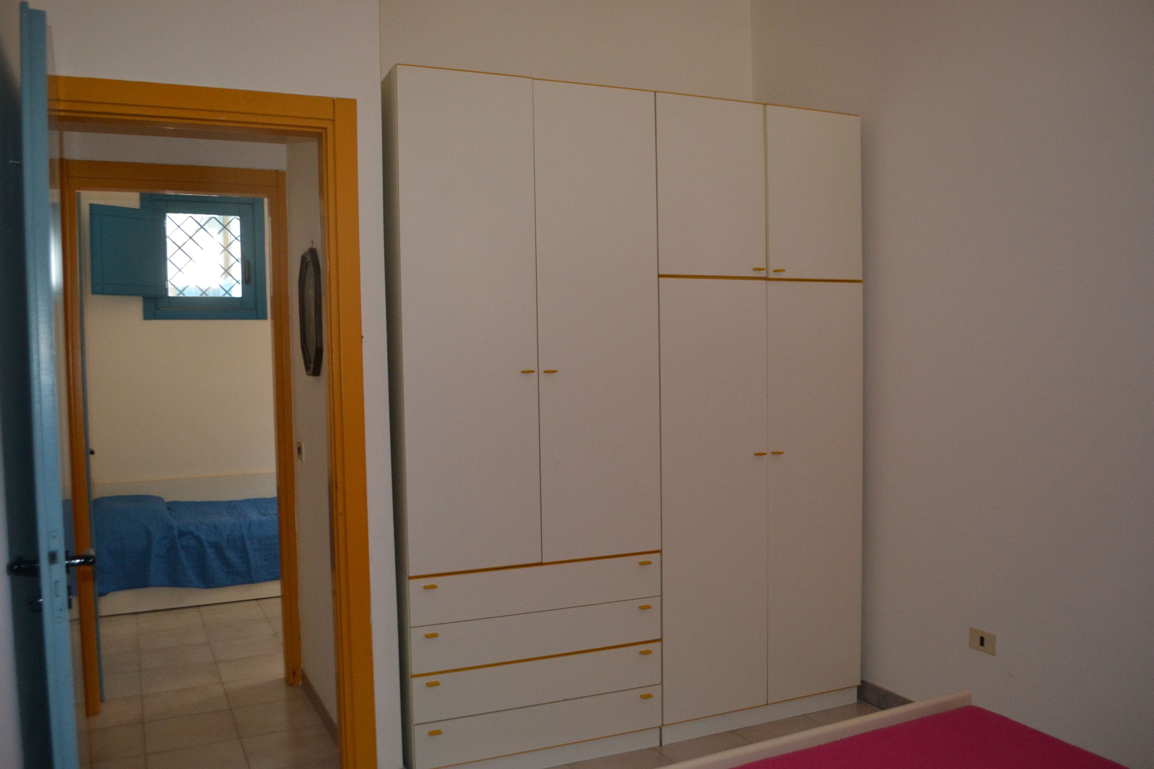 Een kamer met een grote, witte garderobe met meerdere lades en deuren, een bed met een blauwe spreideel, en een raam met blauwe luiken.