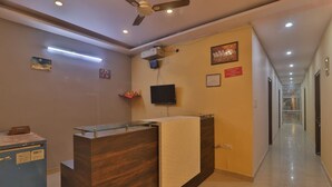 A hotel reception area with a reception desk, a ceiling fan, a flat-screen TV mounted on the wall, and a corridor leading to other rooms. - Hotel Shubhangi Inn (Lucknow)
