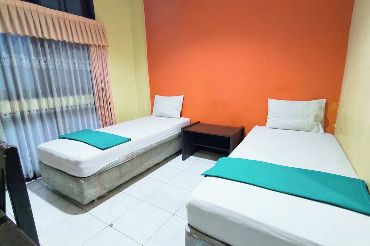 A room with two beds, each with a pillow and a green blanket. There is a small wooden nightstand between the beds.