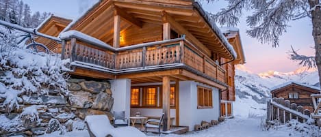 A wooden chalet with a balcony, stone wall, and a snow-covered landscape.