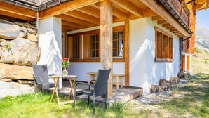 A wooden chalet with a stone wall, two chairs, a table, and a flower vase. - Apartment MARMO by HomeFlow Zermatt (Zermatt)