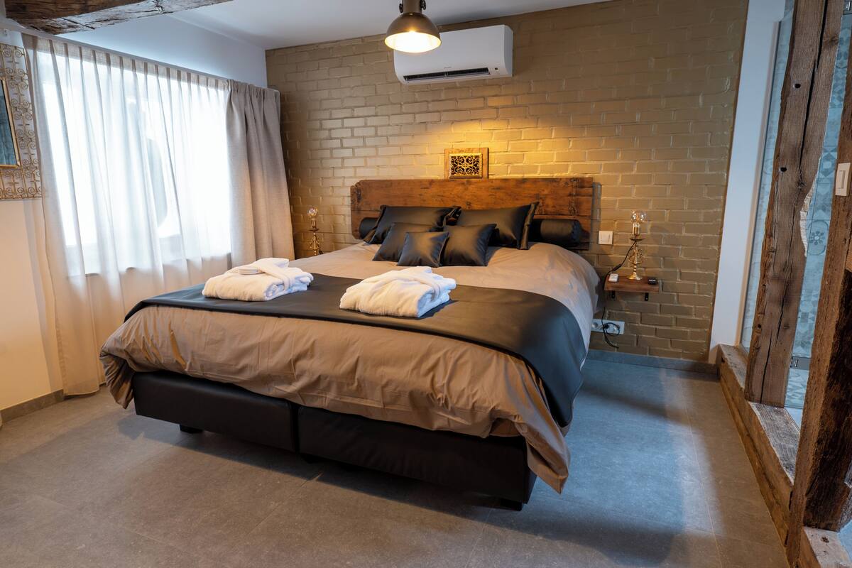 A large bed with a wooden headboard, a bedside table with a lamp, and a window with curtains.