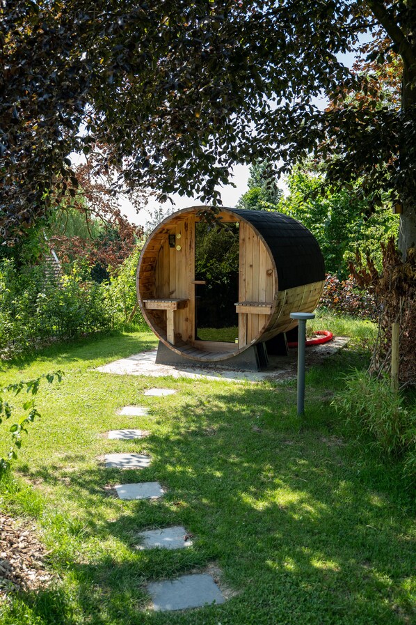 A wooden barrel-shaped structure with a bench inside, surrounded by a garden path and greenery.