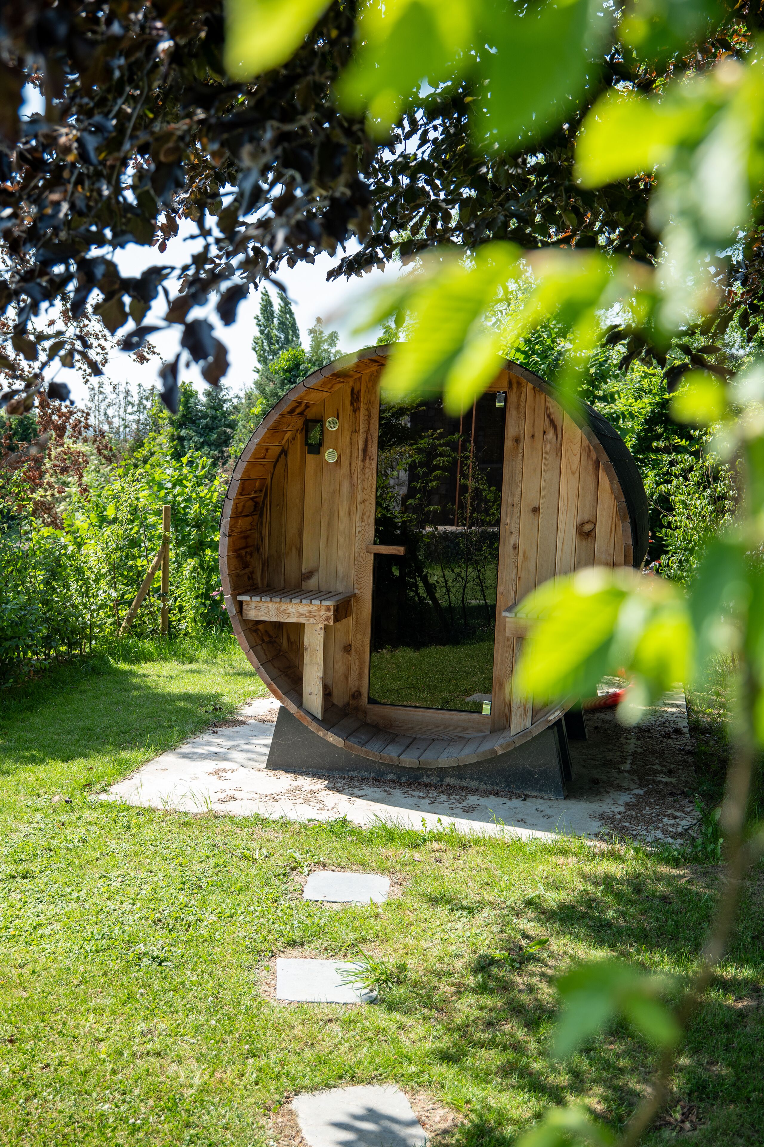 A wooden barrel-shaped structure with a bench inside, surrounded by a grassy area and trees.