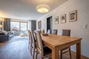A spacious living area with a wooden dining table, comfortable seating, a chandelier, and a large window offering a view of a mountainous landscape. - St. Christoph by HomeFlow (Zermatt)