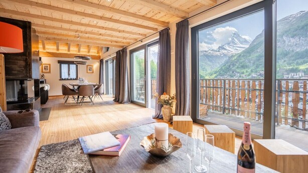 A spacious room with wooden beams, a dining area, and large windows offering a mountain view. - Akelei Penthouse by HomeFlow Zermatt (Zermatt)