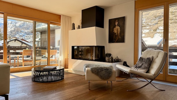 Ein modernes Wohnzimmer mit Kamin, einer bequemen Couch und einem Fernseher. Ein großes Fenster bietet Blick auf eine verschneite Berglandschaft.