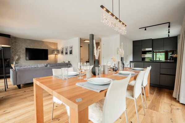 Un espace repas moderne avec une table en bois dressée pour quatre personnes, un canapé gris et une cuisine en arrière-plan.