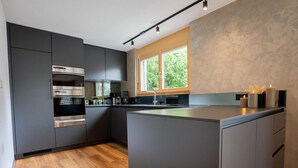 A modern kitchen with dark cabinetry, stainless steel appliances, a large window, and a wooden floor. - Zenith B by HomeFlow Zermatt (Zermatt)