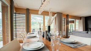 A dining area with a long table set for a meal, large windows with blinds, and a view of the outdoors. - Zenith B by HomeFlow Zermatt (Zermatt)