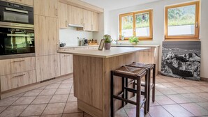 A kitchen with wooden cabinets, a built-in oven, a large island with stools, and a window with a view of greenery. - Galileo by HomeFlow Zermatt (Zermatt)