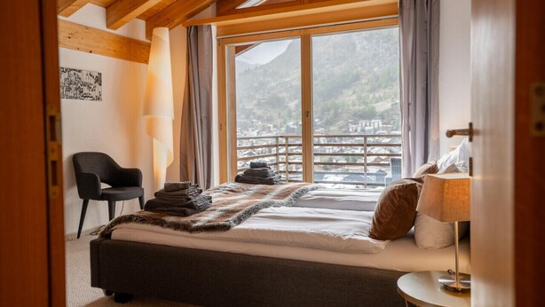 Un dormitorio con una cama, una silla, una lámpara y vistas a un paisaje montañoso a través de amplios ventanales.