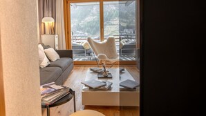 Una sala moderna con un sofá, una silla blanca singular, una mesa de centro con libros y vistas a un paisaje montañoso a través de ventanas amplias.