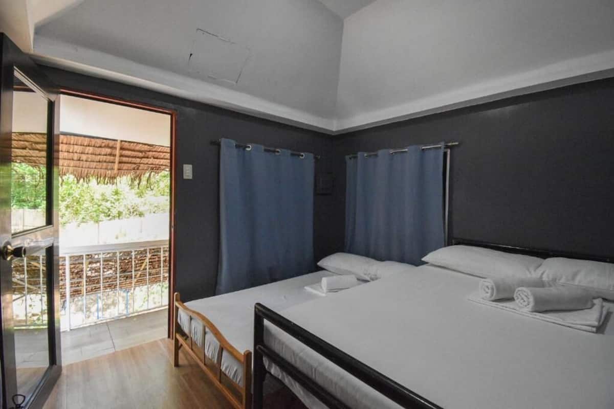 A bedroom with a large bed, two windows with curtains, a balcony with a thatched roof, and a view of greenery.