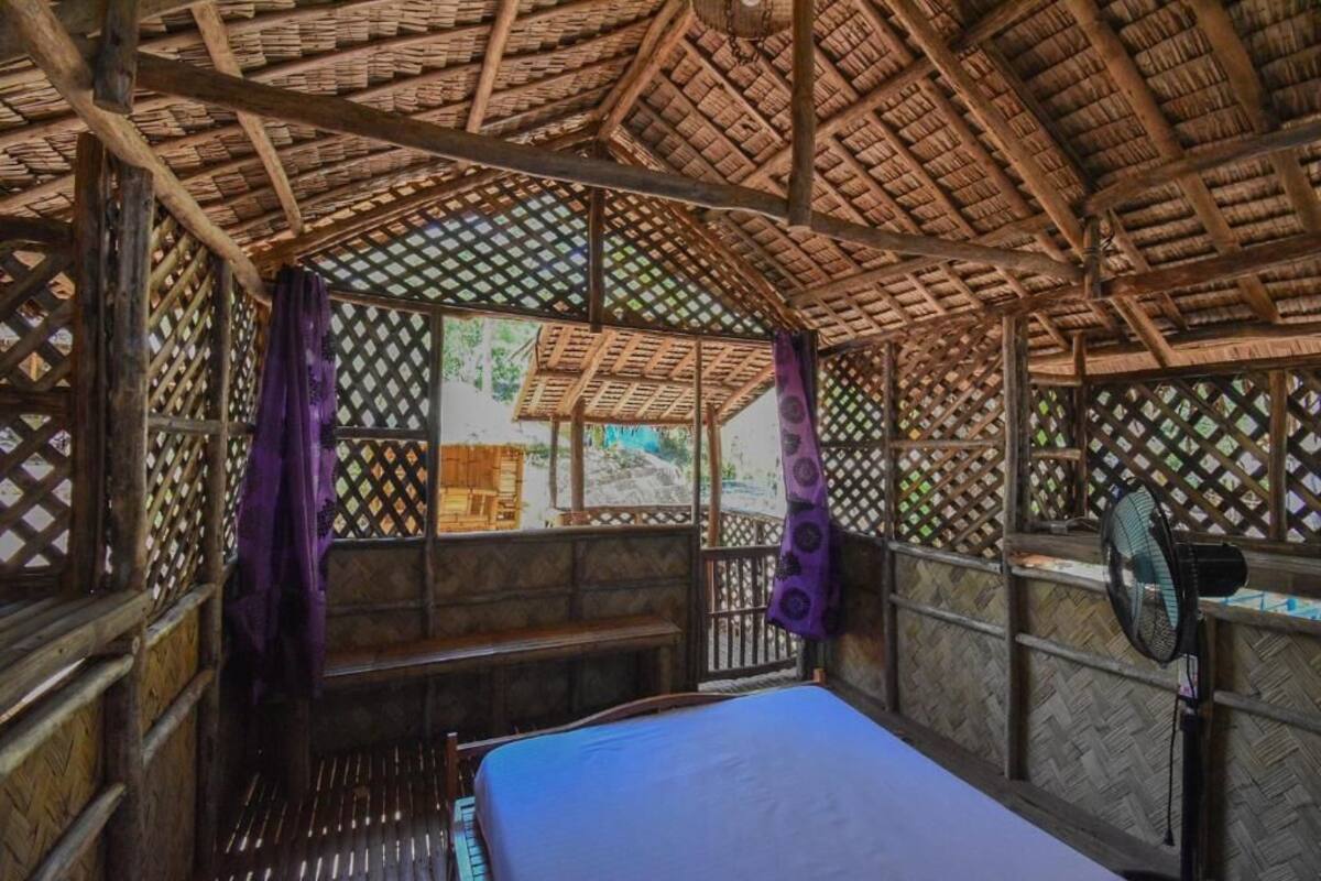 A room with a thatched roof, wooden lattice walls, a bed with a blue mattress, and a fan.