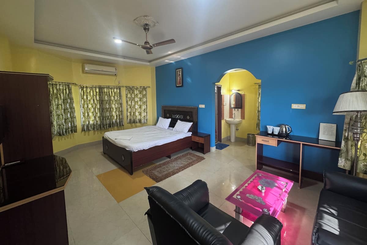 A hotel room with a bed, a desk, a chair, a sofa, a ceiling fan, and a bathroom with a sink and mirror.