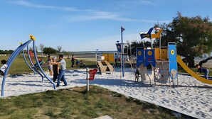 Un parc infantil amb diverses estructures de jocs, incloent tobogans i zones d'escalada, situat sobre un sòl de sorra sota un cel serè.