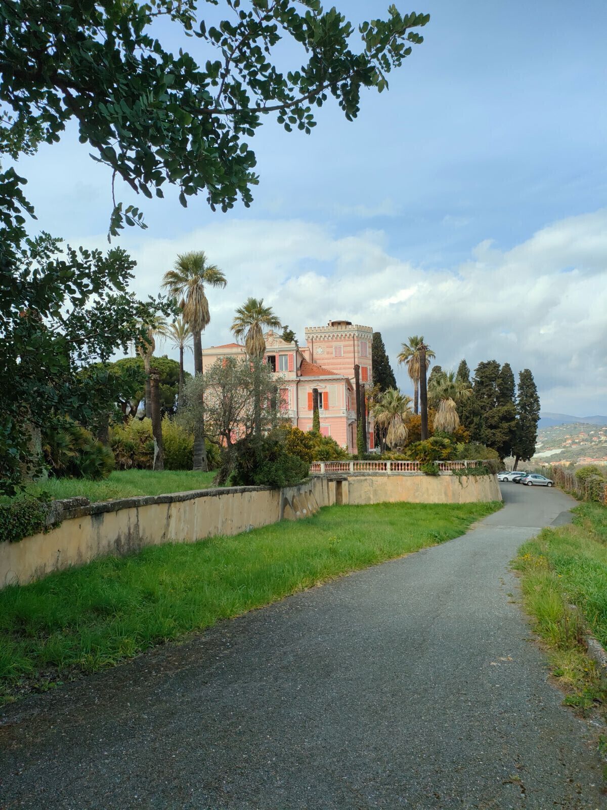 Un chemin pavé mène à un bâtiment rose surmonté d’une tour, entouré de verdure et de palmiers.