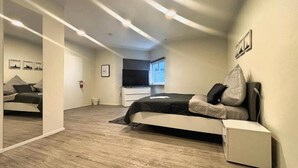 Ein modernes Schlafzimmer mit Bett, Nachttisch und kleinem Schreibtisch. An der Wand befindet sich ein Spiegel und ein Fenster lässt Tageslicht herein.