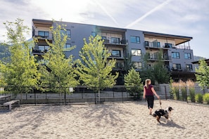 A person walking a dog on a sandy surface with a fenced area, trees, and a multi-story building in the background. - Spacious corner retreat with panoramic mountain views, floor-to-ceiling windows, and designer interiors—minutes from Revelstoke Mountain Resort and downtown. (Revelstoke)