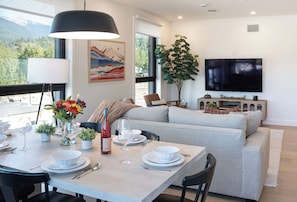 A modern living space with a dining area, sofa, television, and a large window with a view of mountains. - Spacious corner retreat with panoramic mountain views, floor-to-ceiling windows, and designer interiors—minutes from Revelstoke Mountain Resort and downtown. (Revelstoke)