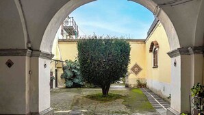 Un couloir en arc de cercle, avec un arbre au centre, un sol pavé de pierres et un bâtiment jaune doté d’une fenêtre et d’une porte.