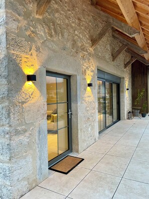 Une façade en pierre, de grandes portes vitrées, une terrasse carrelée et un plafond en bois.