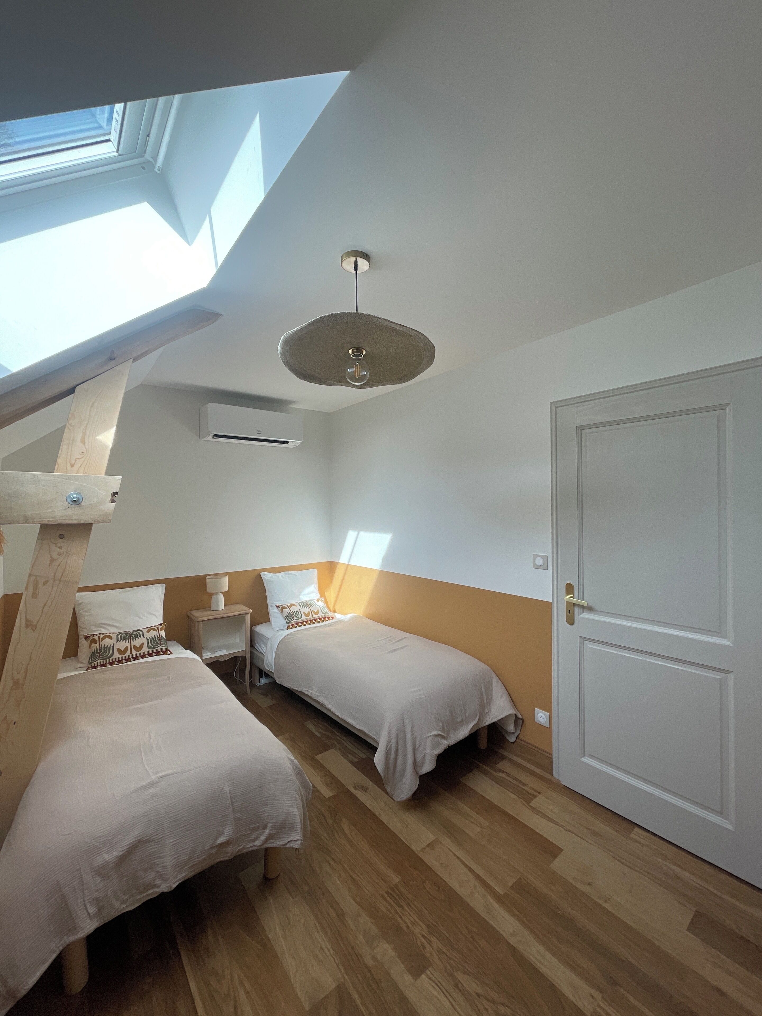 Une chambre avec deux lits, un puits de lumière, un ventilateur de plafond et une porte.