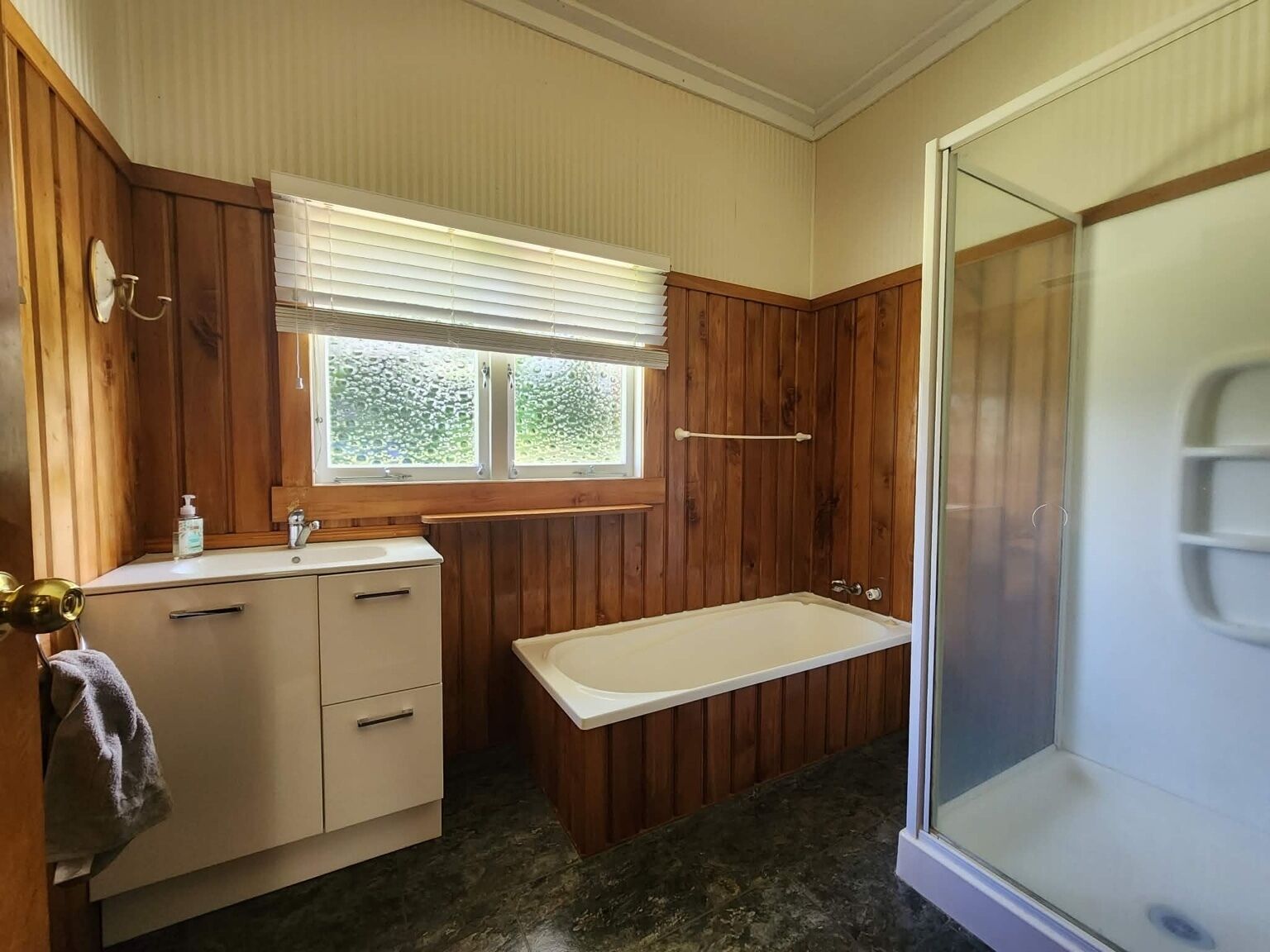 Een badkamer met houten lambrisering, een badkuip, een raam met zonwering en een douchehoek.