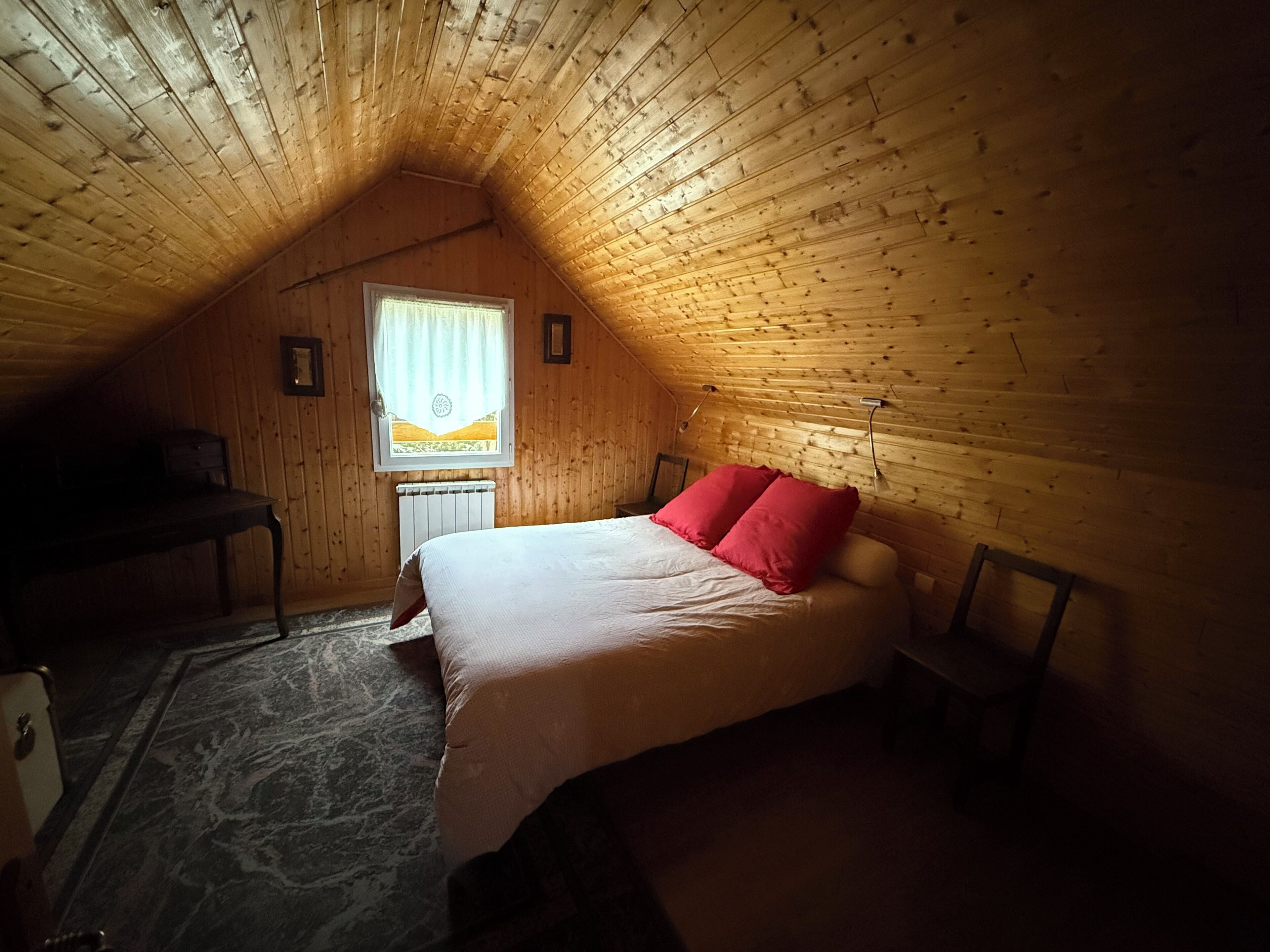 A small, cozy attic bedroom with a sloped ceiling, a single bed with red pillows, a small table, and a radiator.