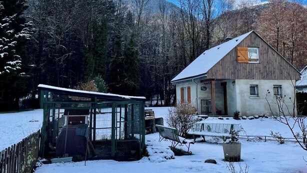 A wooden cabin with a sloped roof, surrounded by a snow-covered landscape and a forested mountain backdrop.