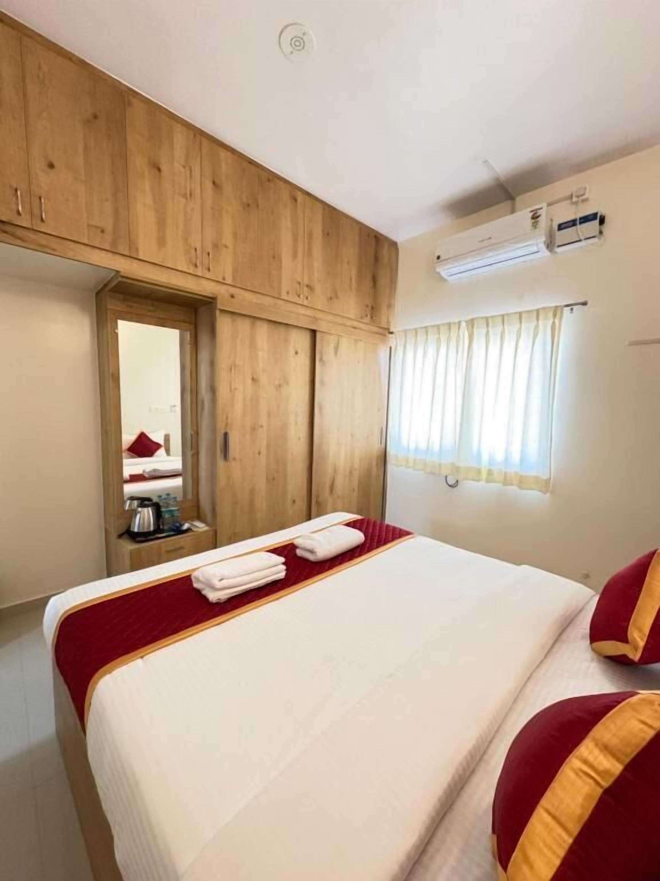 A hotel room with a wooden wardrobe, a bed with white bedding and red pillows, a mirror, and a window with curtains.