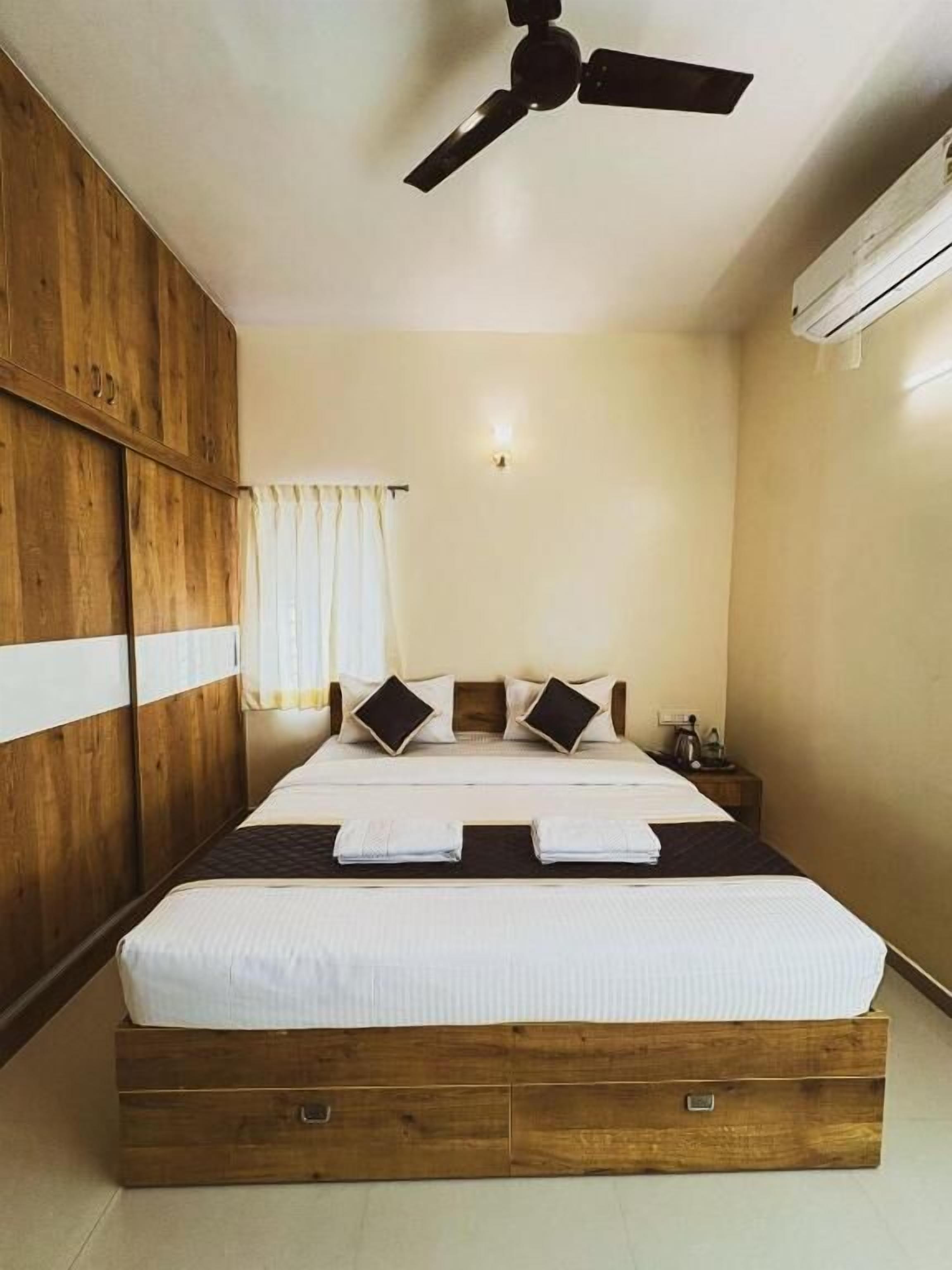 A bedroom with a wooden wardrobe, a bed with white bedding, a ceiling fan, and an air conditioning unit.