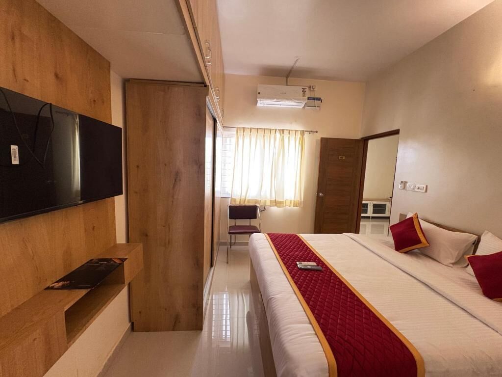 A hotel room with a bed, a TV mounted on the wall, a wooden wardrobe, and a window with curtains.
