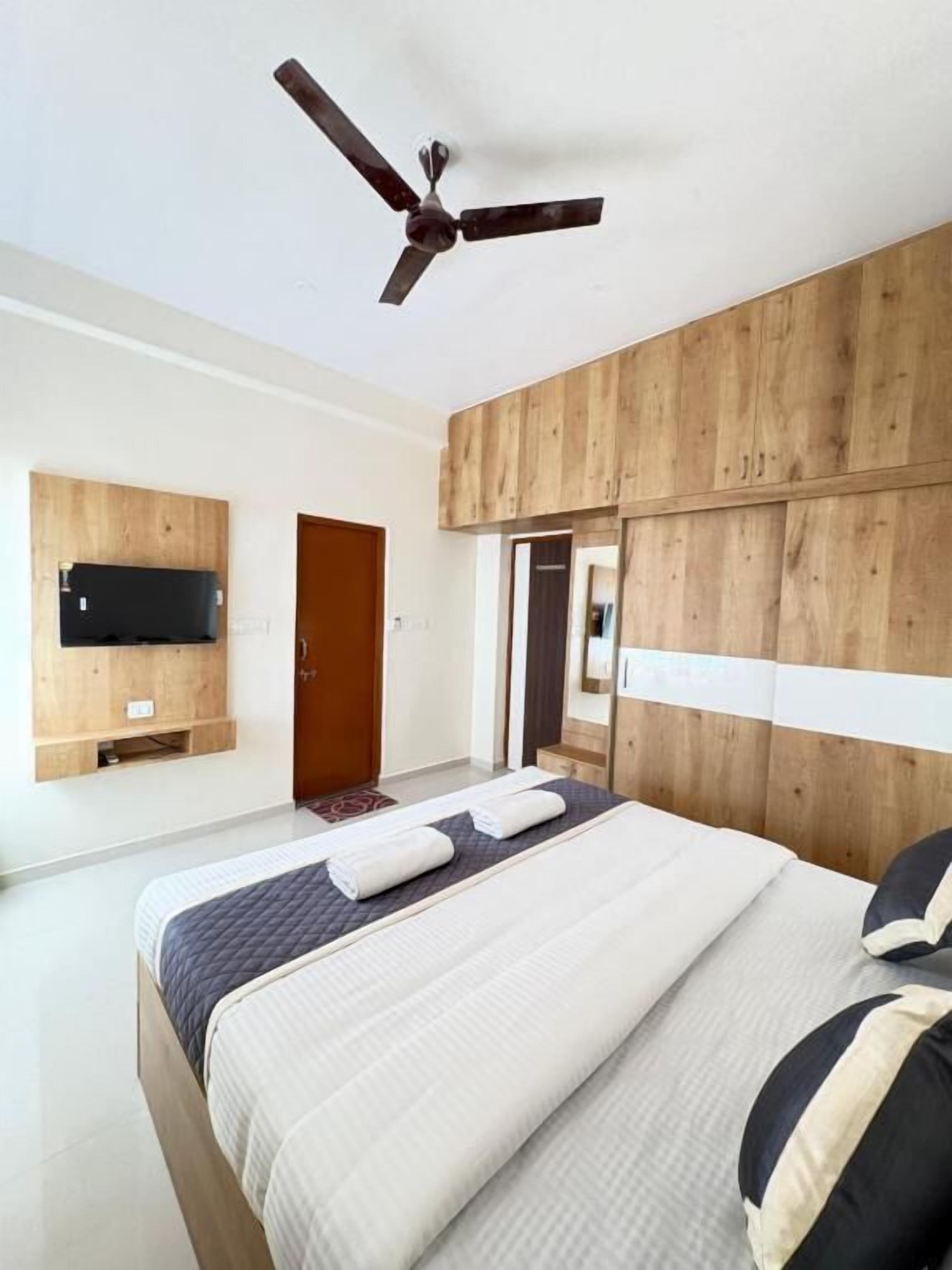 Une chambre à coucher moderne avec une tête de lit en bois, un ventilateur de plafond, un téléviseur à écran plat fixé au mur et un lit bien fait, agrémenté d’oreillers et de serviettes pliées.