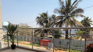 Un balcon doté d’un sol carrelé, d’une rambarde en métal et offrant une vue sur un quartier résidentiel avec des palmiers et un coffret électrique rouge.