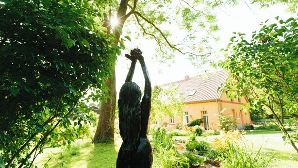 En statue af en person med hænderne løftet i en have med et hus i baggrunden.