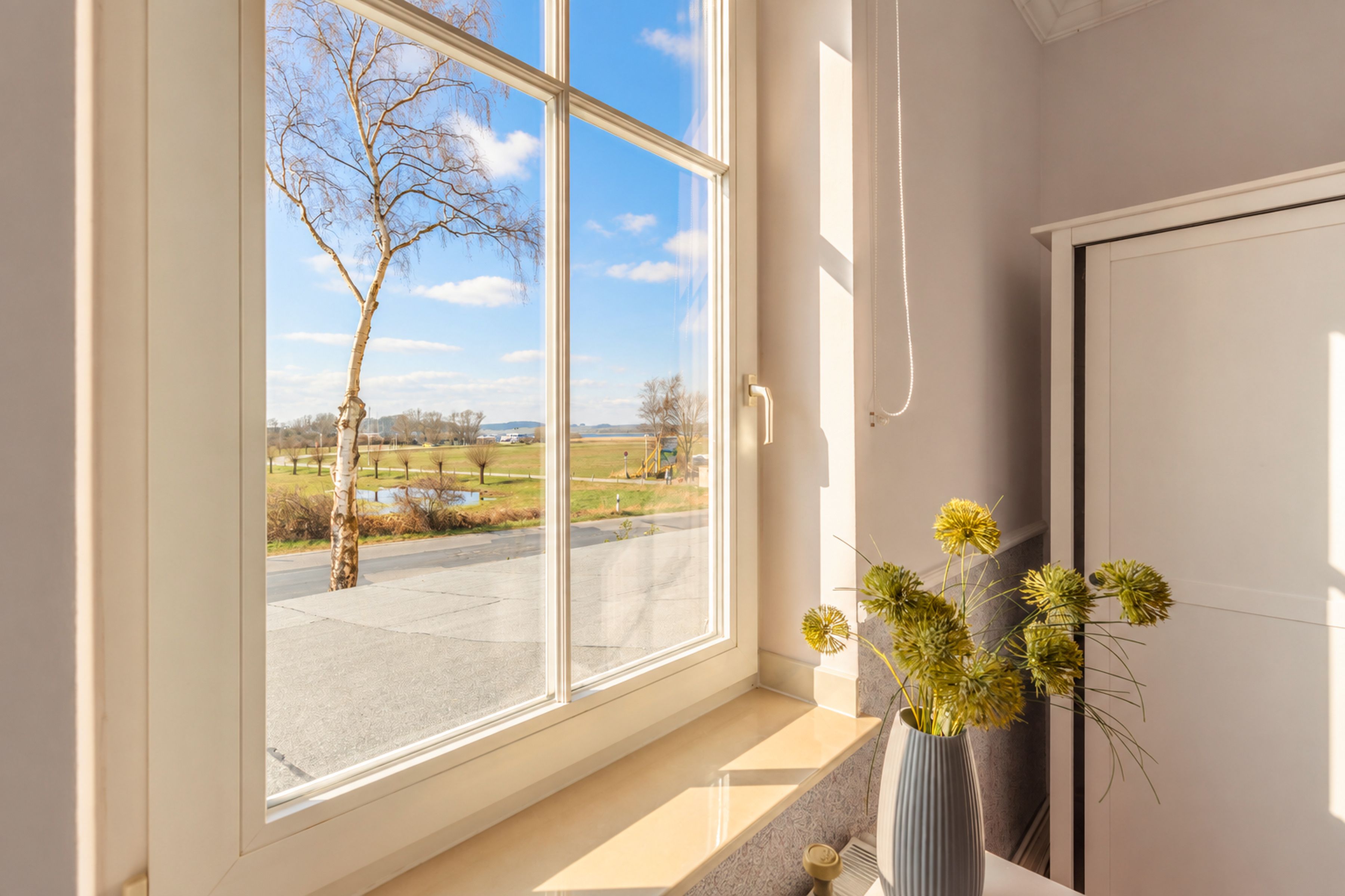 Een kamer met een groot raam, een vaas met gedroogde bloemen en een deur.