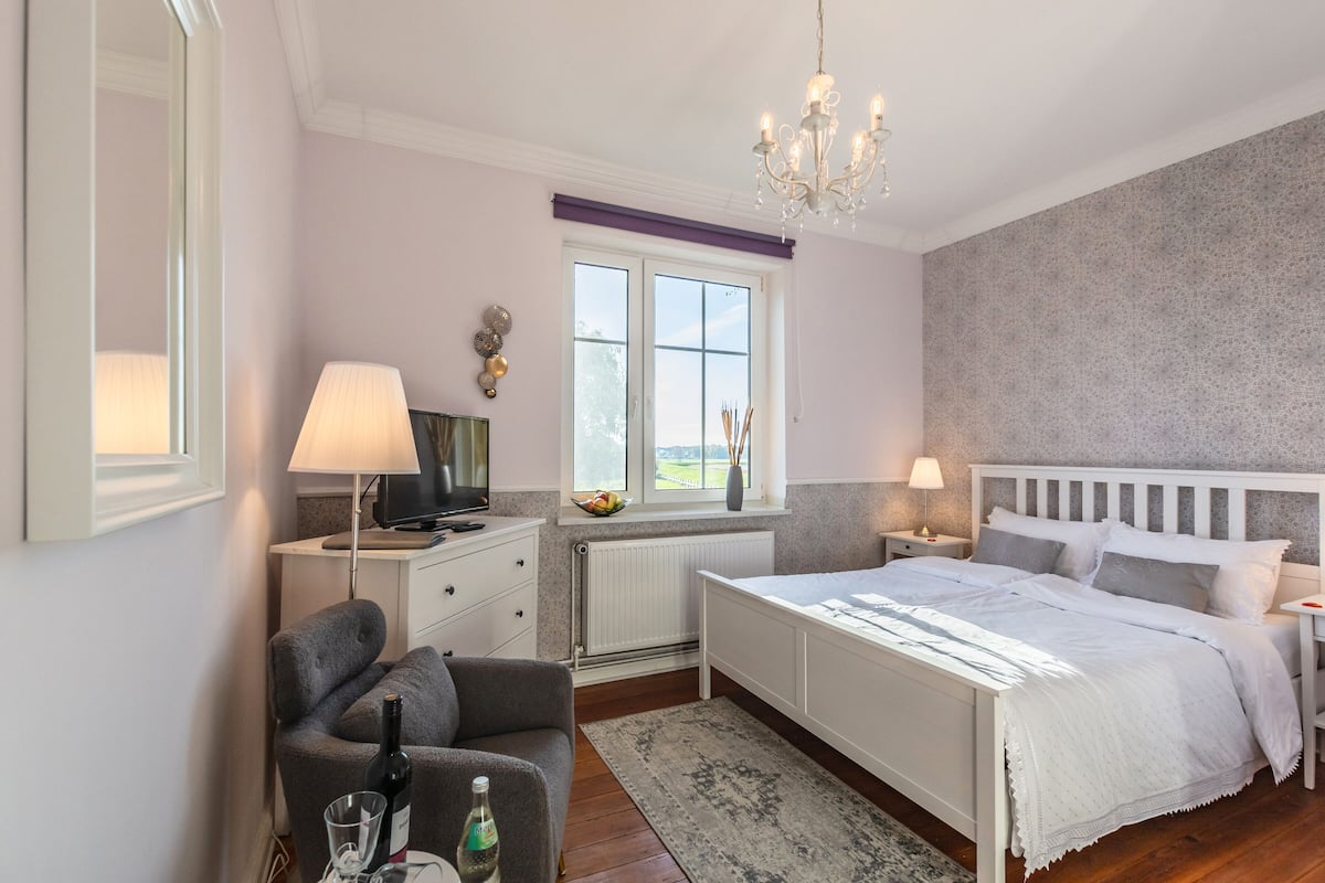 Un dormitorio con cama, una silla, escritorio, lámpara, ventana y candelabro.