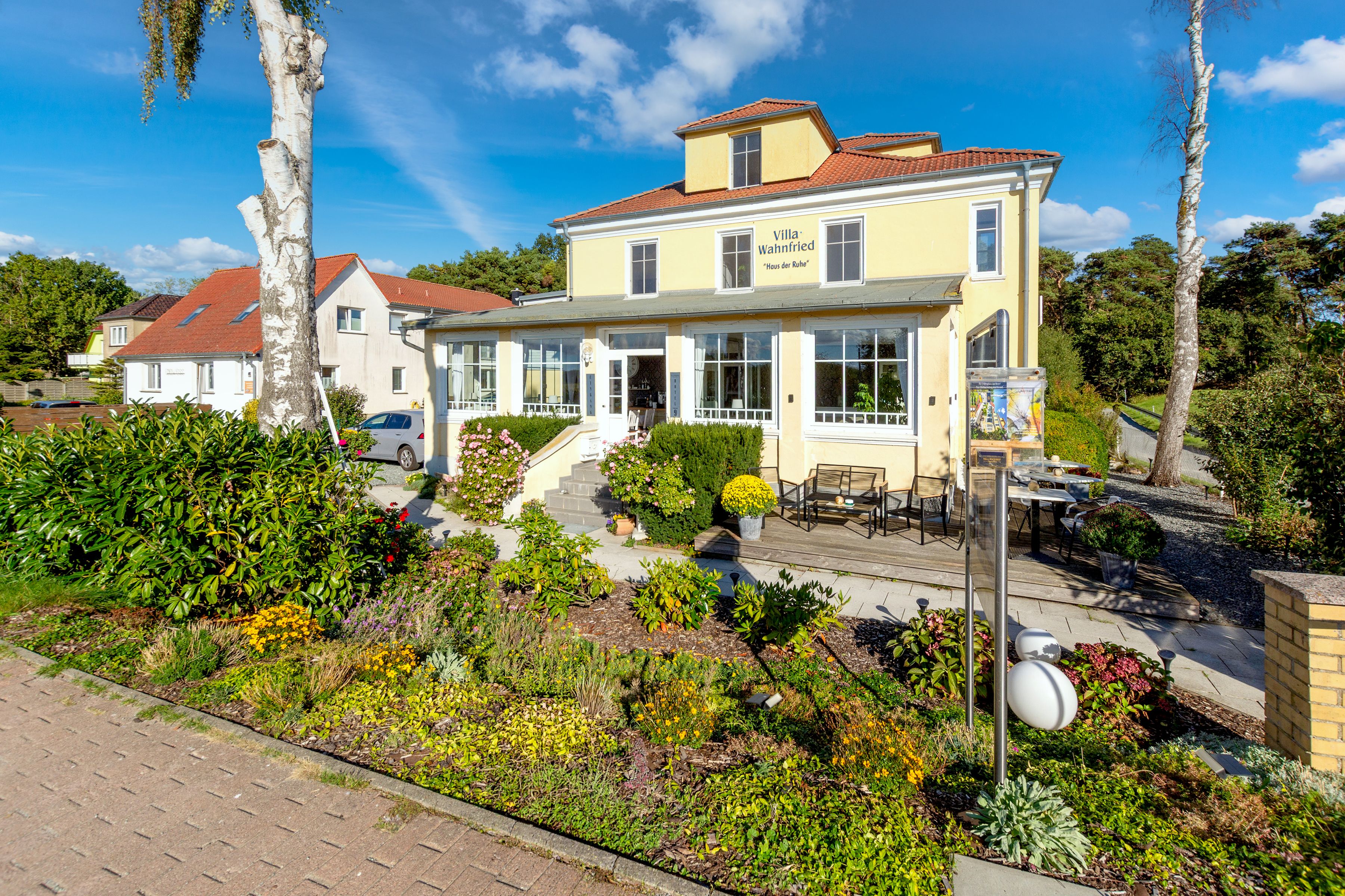 A yellow villa with a red roof, a patio with outdoor furniture, and a garden with various plants and flowers.