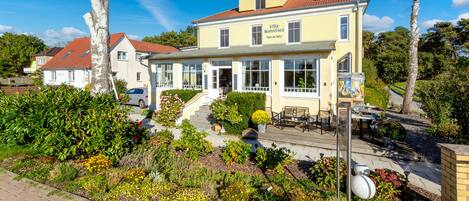 A yellow villa with a red roof, a patio with outdoor furniture, and a garden with various plants and flowers.