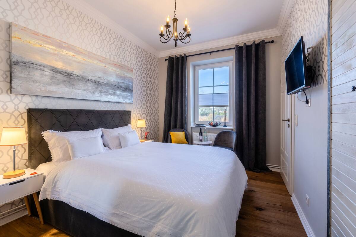 Habitación de hotel con una cama grande, un televisor montado en la pared, una ventana con cortinas y un candelabro.