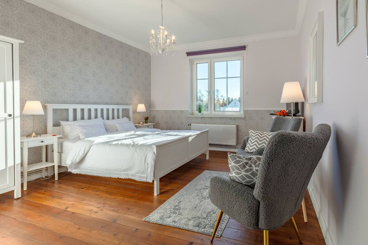 Un dormitorio con cama, mesitas de noche, una silla, una ventana y un candelabro.